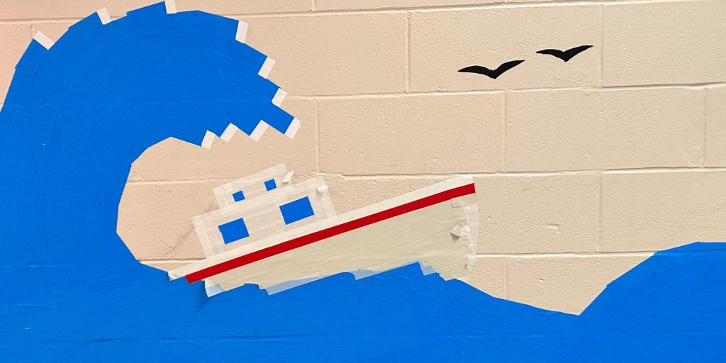 A colorful tape artwork using Pro Tapes products on a brick wall in a school shows a large blue wave engulfing a small red and white ship. Two birds fly overhead. 