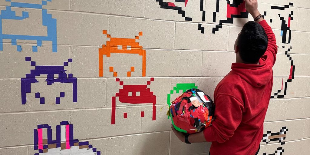 A student removing vibrant tape pixel art from wall and rolling it into a ball.