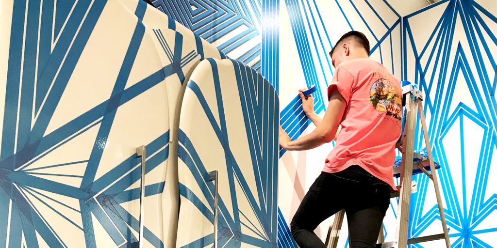 Flekz turned his tape art into a career. We see him up on a ladder, adding new lines to a mural with blue tape.