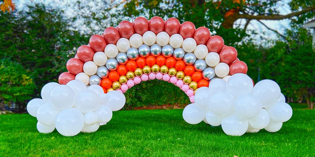 A breathtaking, large rainbow balloon arch by Jexsy Balloons. Featuring red, beige, silver, orange, gold, and pink balloons, it creates a magical scene against a picturesque park backdrop.