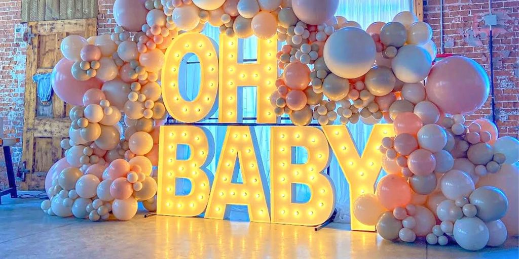 A charming baby shower decoration by Jexsy Balloons. A large pastel pink balloon wall features a warm LED sign that reads 'Oh Baby'.